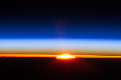 Photographie de l'atmosphère de la Terre prise depuis l'ISS, avec un joli coucher (ou lever ? pas facile à dire sur une photo) au milieu. On voit une bande sombre en haut (l'espace) et en bas (la surface solide de la Terre), et entre les deux, une couche colorée. Elle est rouge-orange au niveau du sol, à cause de la présence du Soleil, bleu sombre près de l'espace où elle se raréfie, et entre les deux, bleu ciel.