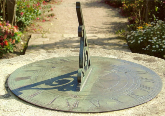 Photographie d'un cadran solaire, trouvée sur la page Wikipédia qui leur est dédiée. On voit une plaque circulaire au sol marquée de chiffres romains de Ⅰ˛à Ⅻ, avec un motif ouvragé dressé qui projette son nombre dessus et fait office d'aiguille. La rotation de la Terre sur elle-même change l'angle par lequel on voit le Soleil, ce qui fait donc avancer l'heure. La révolution de la Terre autour du Soleil fait varier la hauteur avec laquelle on voit celui-ci (on en reparlera plus bas), ce qui joue sur la longueur de cette ombre.