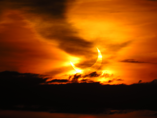 Photo d'une éclipse partielle de Soleil prise aux USA le 10 juin 2021, et trouvée sur Wikimédia Commons. On voit un ciel globalement rouge-orange, avec quelques nuages, et surtout l'image d'un croissant de Soleil, recouvert à plus de la moitié par notre Lune. L'ensemble est assez impressionnant, mais assez loin de ce que c'est que de vivre une éclipse totale.