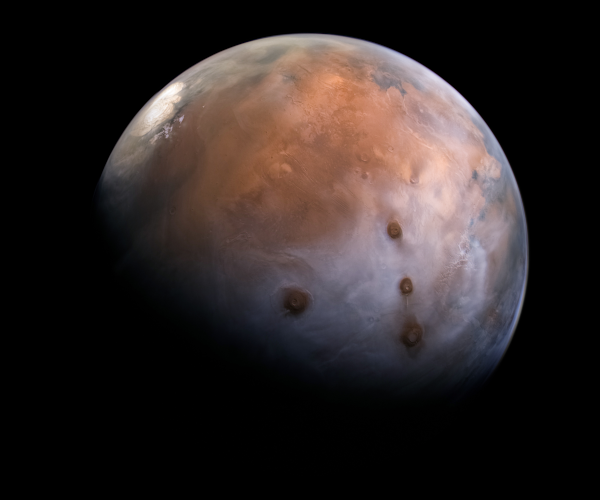 Photographie de Mars (trouvée sur Wikimédia Commons et prise par la sonde Al-Amal, mission martienne lancée par les Émirats arabes unis), montrant simultanément plusieurs des formations les plus impressionnantes de la planète. En haut à gauche, on voit le glacier situé au pôle nord (il y en a un autre au niveau du pôle sud, évidemment invisible ici, mais celui-ci est le plus grand des deux). Tout à droite, dans la moitié haute de l'image, on devine le début de Valles Marineris, un canyon de la largeur des USA. Mais surtout, au milieu de l'image, juste au dessus du terminateur (la limite entre le jour et la nuit sur un corps céleste) on voit le dôme de Tharsis avec ses cinq volcans principaux : à droite les Tharsis Montes, trois volcans alignés (la longueur totale de l'alignement correspond à peu près à celle de la diagonale France-Espagne), et plus à gauche, Olympus Mons, partiellement dans l'ombre, et plus haut Alba Mons, dont on devine le cratère. Ironiquement, l'angle de la photo, le terrain et la météo (on devine quelques nuages par endroits) font d'Alba Mons la partie la plus colorée de l'image.