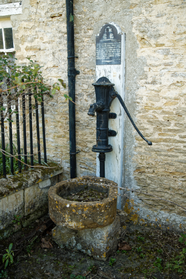 Photo d'une vieille pompe manuelle, trouvée sur Wikimédia Commons. On voit un tuyau métallique servant à faire remonter l'eau fixé au mur, avec une poignée pour pomper et un réservoir en dessous pour recueillir l'eau qui s'écoule. Une plaque gravée, illisible sur cette image, donne quelques informations de contexte au dessus. En actionnant le levier, on vient chasser l'air dans le tuyau. On pensait jusqu'à l'époque de Galilée que c'était ce qui aspirait l'eau vers le haut, pour empêcher du vide de se former ; on comprendra bientôt que c'est en fait l'air qui, de l'autre côté du tuyau, pousse l'eau et la fait monter aussi haut que possible tant que rien ne vient la bloquer. Une fois l'eau montée jusqu'au dessus du tuyau de sortie, c'est simplement la gravité qui la fait retomber du bon côté, de la même manière que c'est essentiellement la gravité qui la fait avancer dans les aqueducs romains, qui sont toujours plus ou moins penchés.