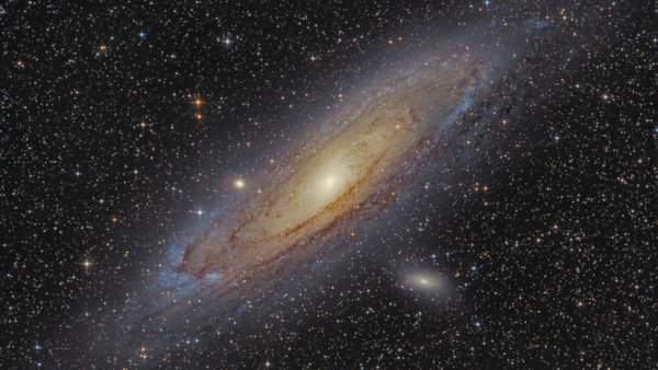 Photo, sur fond de ciel étoilé, de la galaxie d'Andromède telle qu'on peut l'admirer dans notre ciel au télescope. Il s'agit d'une galaxie spirale ressemblant vaguement à la nôtre, mais vue avec un angle qui nous la présente un peu de côté. Comme toutes les galaxies, on voit qu'elle a des couleurs différentes selon les concentrations d'étoiles et de gaz (elle tire plus sur le jaune vers le centre et sur le bleu à sa périphérie), et que la partie centrale est plus lumineuse, car constituée d'un grand nombre d'étoiles qui semble assez proches les unes des autres vues de cette distance.