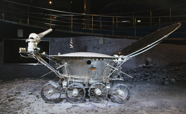 Reproduction du Lunokhod 1 exposée au musée de l'astronautique à Moscou, et trouvée sur la page Wikipédia anglophone dédiée au Rover. On voit ce qui ressemble à une surface de gravas en bas avec des rampes pour le public en arrière-plan, et bien sûr, occupant la plus grosse partie de l'image, l'engin, en forme globalement de plus ou moins cylindre avec un bras équipé de divers équipements d'un côté, et de l'autre, ce qui ressemble à un couvercle ouvert. En bas, le robot est équipé de quatre paires de roues l'une derrière l'autre.
