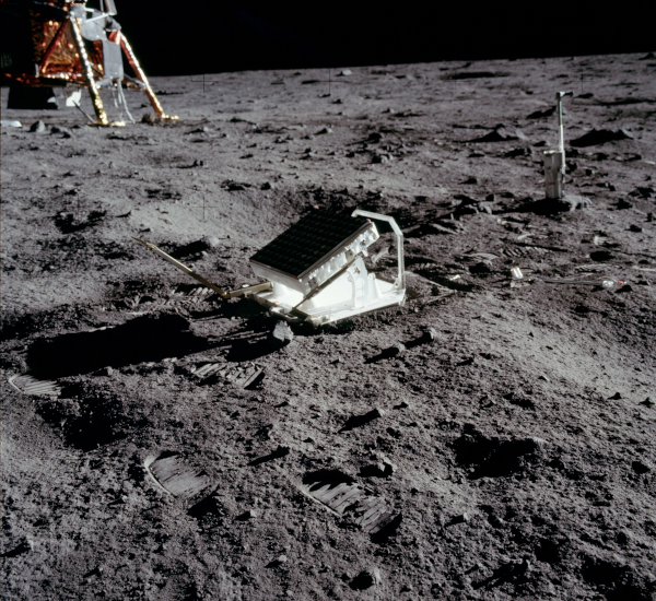 Photo du réflecteur lunaire déposé par l'équipage d'Apollo 11, trouvée sur la page Wikipédia sur les réflecteurs lunaires. On voit la surface lunaire, grise et globalement couverte de poussière, avec quelques empruntes de pas, et au milieu, l'objet à la surface carrée, posé légèrement incliné pour être bien en direction de la Terre. Une partie du LEM est visible en arrière-plan dans le coin en haut à gauche.