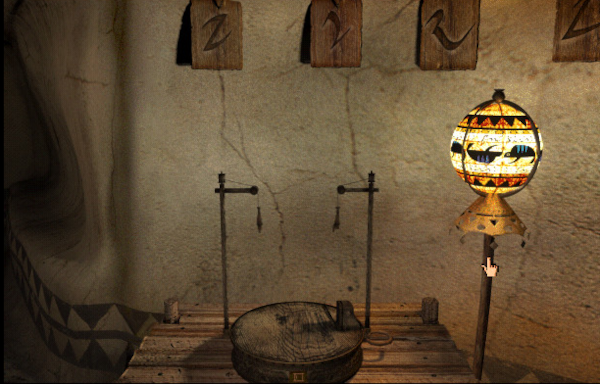 Nouvelle capture d'écran de Riven, montrant cette fois-ci la salle de classe, et plus particulièrement le jouet en bois permettant d'apprendre à compter. Il s'agit d'un objet de bois dans lequel deux petites figurines représentant des personnages sont pendues par les pieds au dessus d'un plateau sur lequel la tête d'un animal marin est représentée bouche ouverte. Chaque fois qu'on enclenche le mécanisme, l'une des deux figurines descend d'un certain nombre de crans, facile à compter, et le symbole correspondant est affiché en écriture rivenienne en bas du plateau, ce qui nous permet d'associer ce symbole au nombre. À chaque activation, la tête de poisson change de côté, et la première figurine qui descend jusqu'à toucher le plateau se fait donc « manger », j'imagine qu'il est censé s'agir d'une sorte de petit jeu aléatoire pour les élèves. À part ça, la scène est éclairée par une petite lampe décorée de motifs triangulaires et de formes d'animaux marins, et on peut voir accrochées au mur les pages d'un abécédaire montrant que les gens qui vivent ici ont aussi un alphabet différent du nôtre.