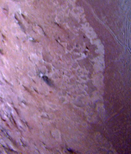 Photographie, prise par Voyager 2 et trouvée sur Wikimédia Commons, d'un cryovolcan identifié sur Triton. La photo est en « fausses couleurs », c'est-à-dire qu'il s'agit d'une composition de trois photos en noir et blanc (deux prises en lumière visible, à deux fréquences différentes, et une prise dans les ultraviolets) qui ont été ramenées sur les canaux rouge, vert et bleu pour nous permettre de voir davantage de détails à la fois. On a donc l'impression de voir un sol globalement violet, mais ça ne correspond pas aux couleurs naturelles de cette lune. On voit ici globalement une portion rectangulaire de la surface avec environ au centre (légèrement sur la gauche) une zone plus sombre qui correspond à un cratère avec une coulée.