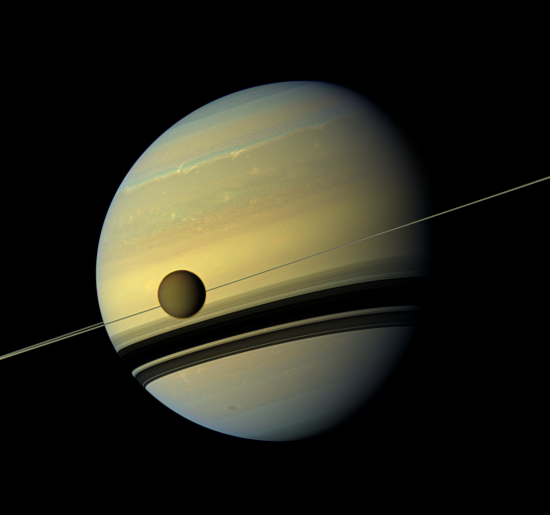 Photographie de Titan devant Saturne, prise par la sonde Cassini et trouvée sur Wikimédia Commons. On voit la planète géante avec ses anneaux quasiment vus par la tranche, donc très fins, et légèrement de biais, même si on distingue leurs ombres beaucoup plus grandes sur la planète. Au premier plan, une lune passe pile devant ces anneaux, elle est d'une couleur assez proche de celle de la planète, mais beaucoup plus sombre.