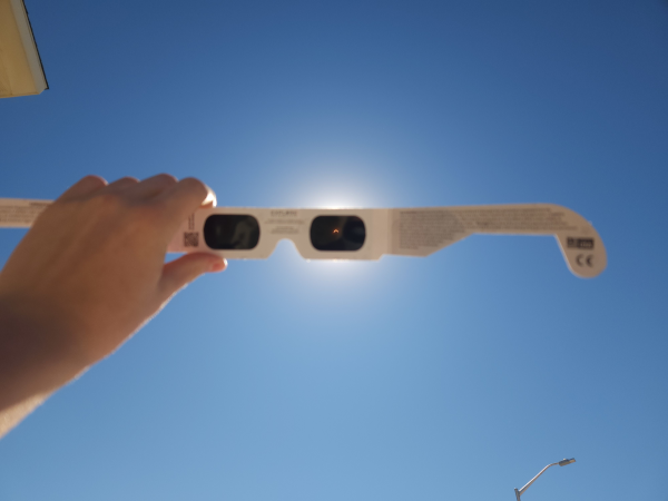 Lunettes d'observations d'éclipse, photo trouvée sur Wikimédia Commons. On voit un ciel bleu (avec un lampadaire en bas et le coin d'un toit en haut, mais on peut les oublier) devant lequel une main tient la paire de lunettes en question, d'un modèle assez semblable à celui qui illustrait le pouet d'introduction. On a donc le “verre” de gauche, toujours noir, mais on voit cette fois le Soleil au travers du “verre” de droite (on remarque d'ailleurs le halo lumineux autour des lunettes à ce niveau). C'est bien sûr tout petit (on parle de comment agrandir ça juste en dessous), mais on peut quand même distinguer que l'éclipse est bien avancée, la Lune nous masquant une bonne partie de notre étoile et ne laissant voir qu'un petit croissant de Soleil… qui éclaire quand même pas mal à lui tout seul, puisque la scène n'a pas l'air spécialement plus sombre que d'habitude.