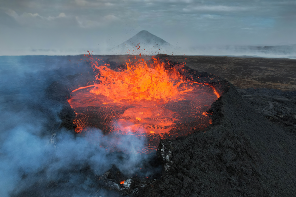 Photo, trouvée sur Wikipédia, d'une éruption volcanique à Fagradalsfjall en Islande, en 2023. L'image a été prise par un drone, permettant de s'approcher davantage du cratère que ce qui serait raisonnable pour des êtres humains. On voit une montagne à l'arrière-plan, et au premier, le sommet du cratère avec une étendue bouillonnante de lave rouge, dont s'échappe aussi pas mal de fumée.