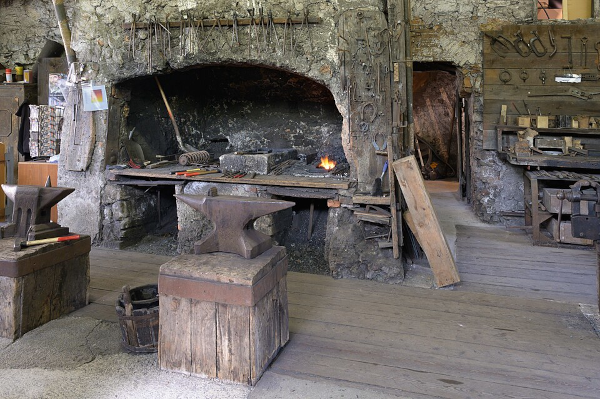 Photo de l'intérieur d'une forge, trouvée sur Wikimédia Commons. On voit une large cheminée de pierre au mur de laquelle plusieurs pinces sont accrochées, et au premier plan, une enclume posée sur un billot. Le forgeron, absent de l'image, utilise le feu pour chauffer au rouge les pièces de métal sur lesquels il travaille, ce qui lui permet de les ramollir pour pouvoir ensuite leur donner la forme dont il a besoin avec son marteau.