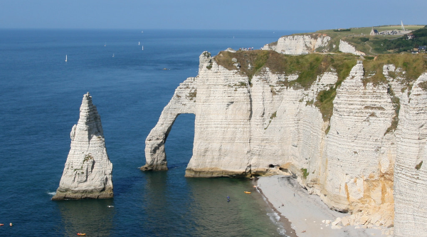 Photo de la falaise d'Étretat, avec une plage à droite et la célèbre aiguille, théâtre d'aventures d'Arsène Lupin, dans l'eau sur la droite. Entre les deux, au centre de l'image, la falaise s'avance dans la mer, et on peut voir une arche de pierre en dépasser, et qui peut, avec des reliefs marquants les yeux et les oreilles, donner l'impression d'une tête d'éléphant d'Asie dont la trompe, un peu trop épaisse, plongerait dans l'eau.