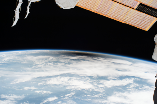 Photographie prise depuis l'espace de l'éclipse du 21 août 2017, trouvée sur Wikimédia Commons. On voit notre planète en bas avec sa couverture de nuages, et en haut, l'espace, ainsi qu'une partie des panneaux solaires de l'ISS au premier plan. Au centre de l'image, en dessous de l'atmosphère (qui continue de diffuser la lumière comme on en a parlé la semaine dernière), une tache sombre couvre une partie de notre planète : il s'agit de l'ombre de la Lune, et ce sont donc les gens situés dans cette zone qui ont pu admirer l'éclipse totale.
