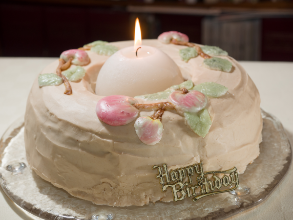 Photo d'un gâteau d'anniversaire trouvé sur Wikimédia Commons. Il s'agirait apparemment d'un gâteau aux pommes avec quelques petites décorations dessus et un petit « Happy Birthday » posé devant… et surtout, le gâteau ayant une forme globale de donut, une grosse bougie allumée dans le trou au centre.