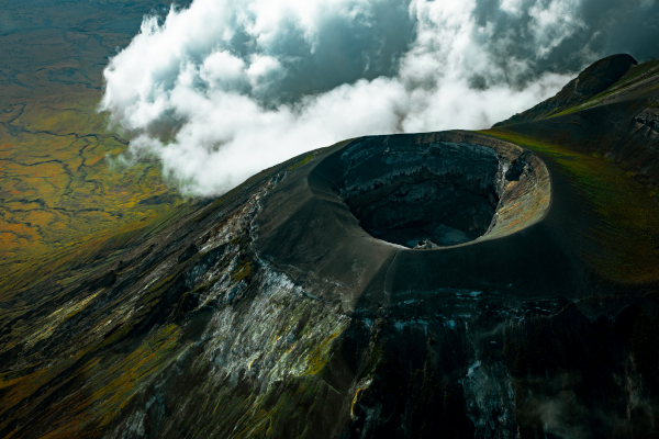 Vue aérienne du volcan, trouvée sur Wikimédia Commons. On voit la plaine avec des nuages en contrebas, et surtout le sommet du volcan, avec un cratère à peu près circulaire à l'intérieur duquel on aperçoit la surface d'un liquide noir.