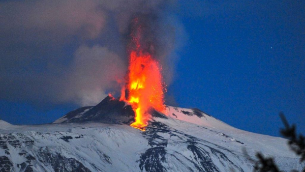 Photo d'une éruption de l'Etna, un volcan de type strombolien situé en Italie. On voit la montagne couverte de neige, et au centre, la lumière rouge de la lave, qui à la fois commence à couler le long du volcan comme dans une éruption effusive, et à la fois est projetée vers le ciel avec pas mal de fumée comme dans une éruption explosive.