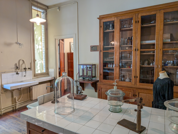 Photo de la reconstitution du laboratoire de chimie de Marie Curie, présent au musée Curie (et trouvée sur Wikimédia Commons). C'est évidemment un labo bien plus moderne que celui de Torricelli, étant donné la différence d'époque, mais on y voit un robinet, un placard au fond contenant divers produits, et surtout, posés sur une table, quelques outils, notamment des cloches, pouvant servir à contrôler précisément les conditions d'une expérience (la pression de l'air, par exemple, puisque c'est de ça qu'on parle ici). Ça n'empêche pas ces expériences d'être dangereuses (on en reparlera une autre fois, mais Marie Curie est morte de son exposition aux produits radioactifs qu'elle manipulait), mais ce n'est évidemment pas pareil que de laisser les choses se faire dans le monde chaotique autour de nous où toute chose n'est jamais égale par ailleurs.