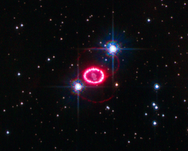 Rémanent de la supernova de 1987. On voit, sur fond de ciel étoilé (deux de ces étoiles sont plus lumineuses que les autres car elles sont situées plus près de nous que l'objet) un anneau rose constitué par les couches de gaz soufflées lors de l'explosion. Autour de lui, des lignes roses plus sombres indiquent que de la matière avait déjà été expulsée vers l'espace lorsque celle-ci s'est produite.